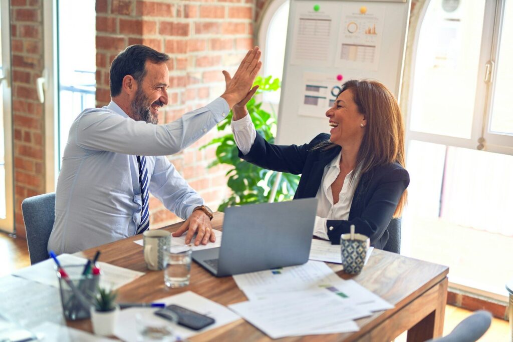 Two colleagues joyfully high-five over a desk cluttered with documents and a laptop, celebrating teamwork in a bright office setting.