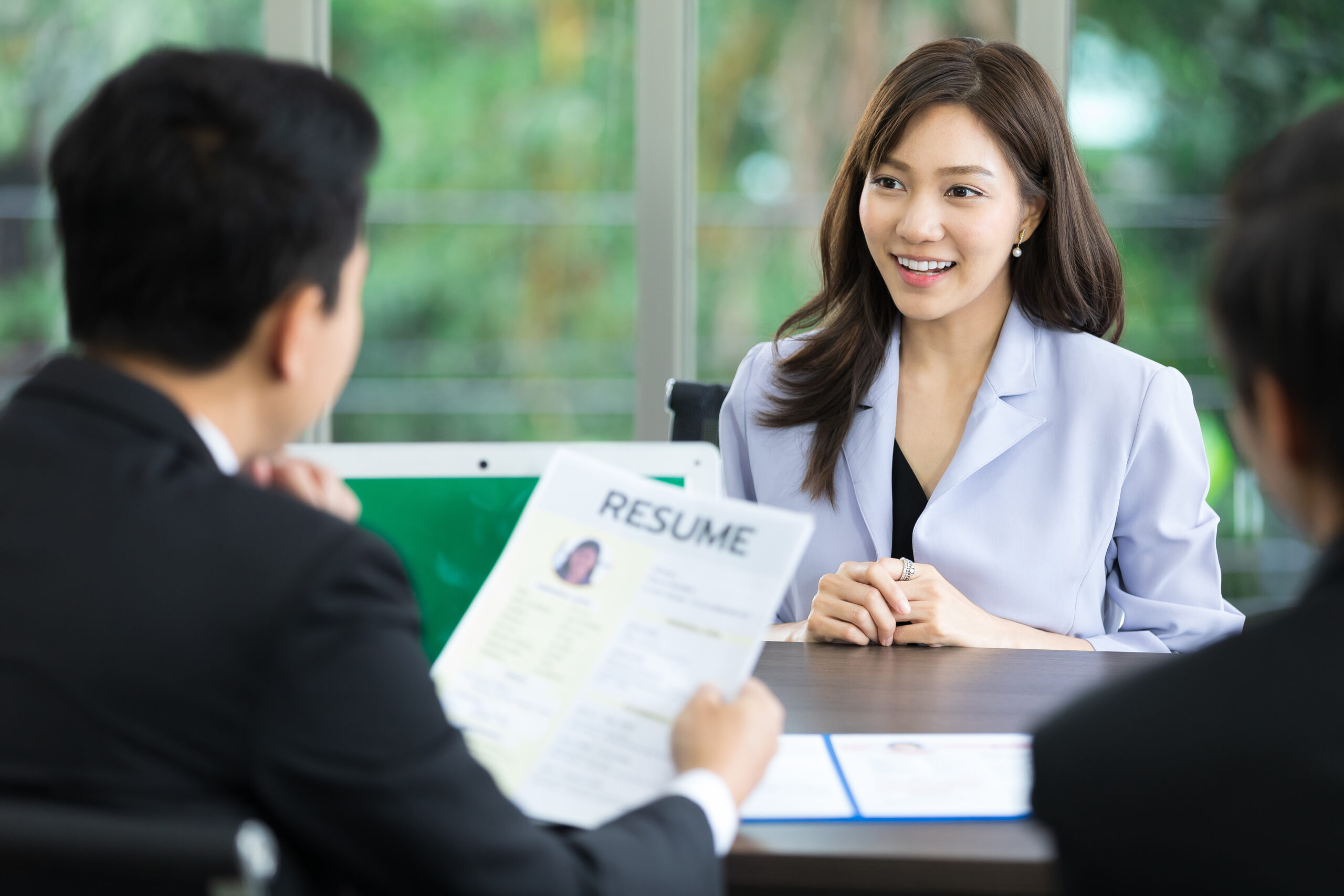 A woman in a light blazer smiles during a job interview as a man reviews her resume, highlighting a professional setting focused on hiring.