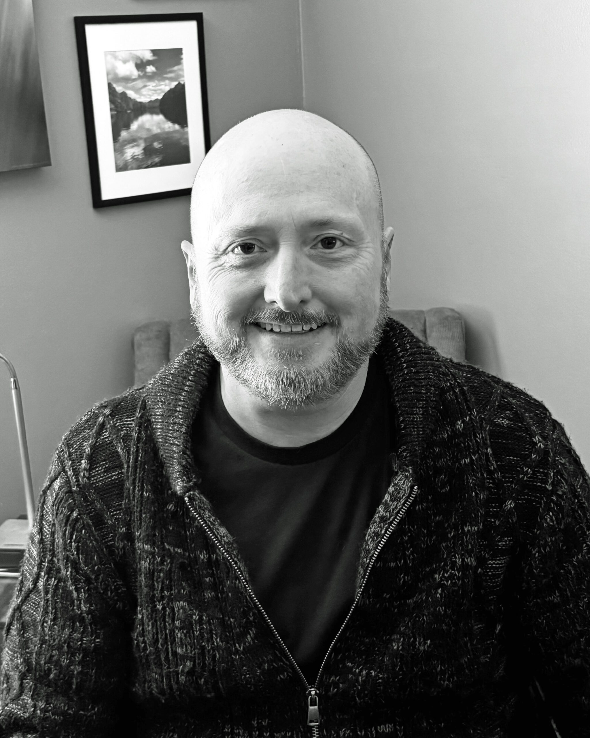 A smiling man with a bald head and a beard wears a dark sweater. A framed landscape photo is visible in the background, adding context.