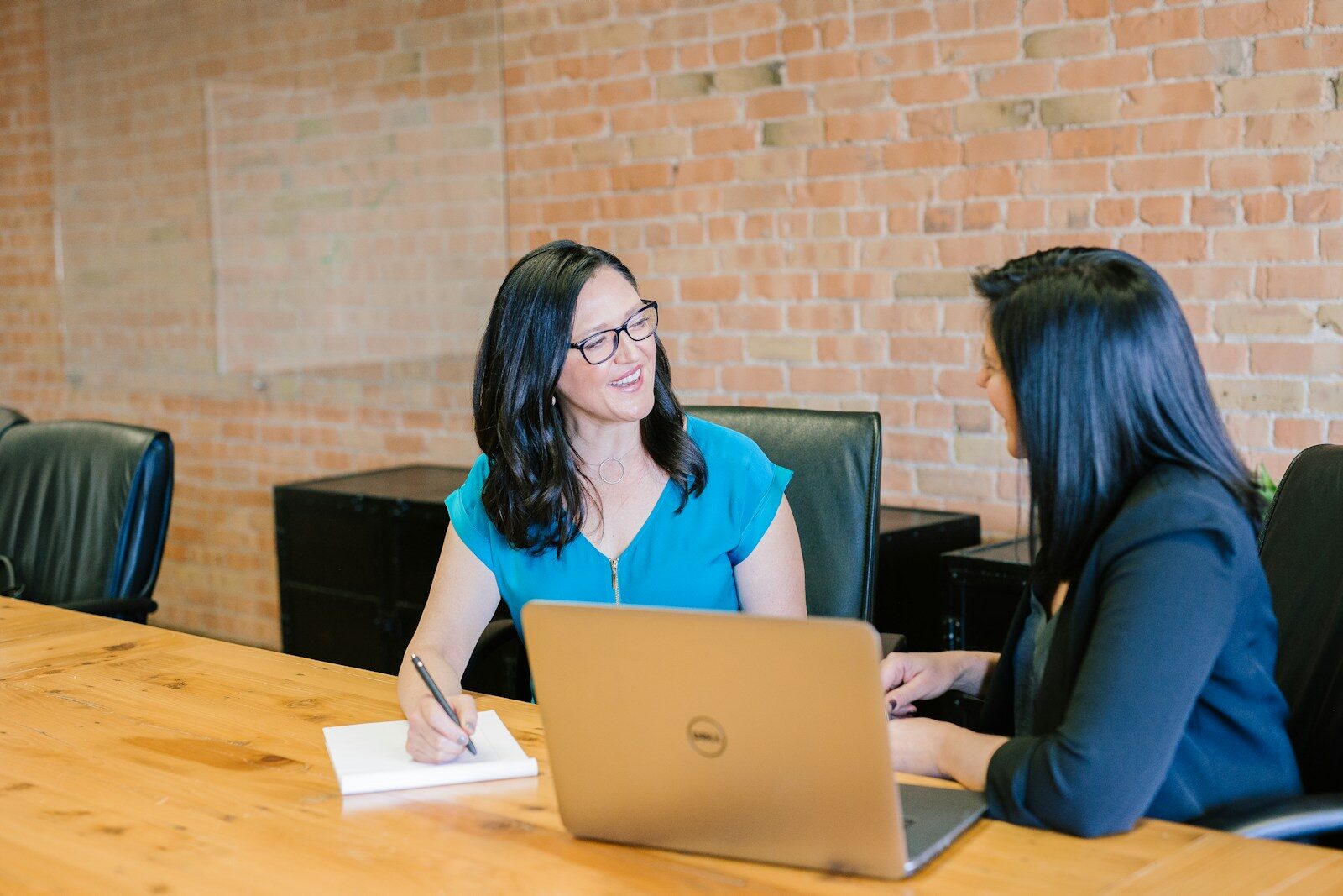 Two women engage in a discussion at a wooden conference table, with one taking notes and the other smiling, highlighting a collaborative work environment.