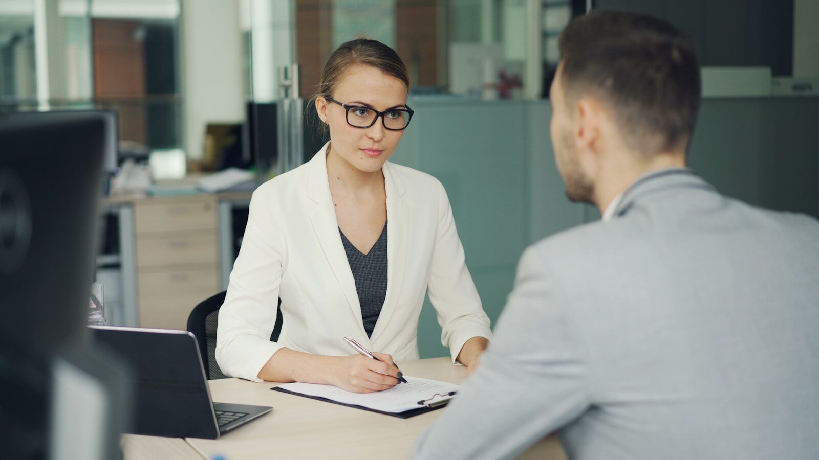A businesswoman in a white blazer takes notes during an interview with a man in a gray suit, highlighting a professional setting.